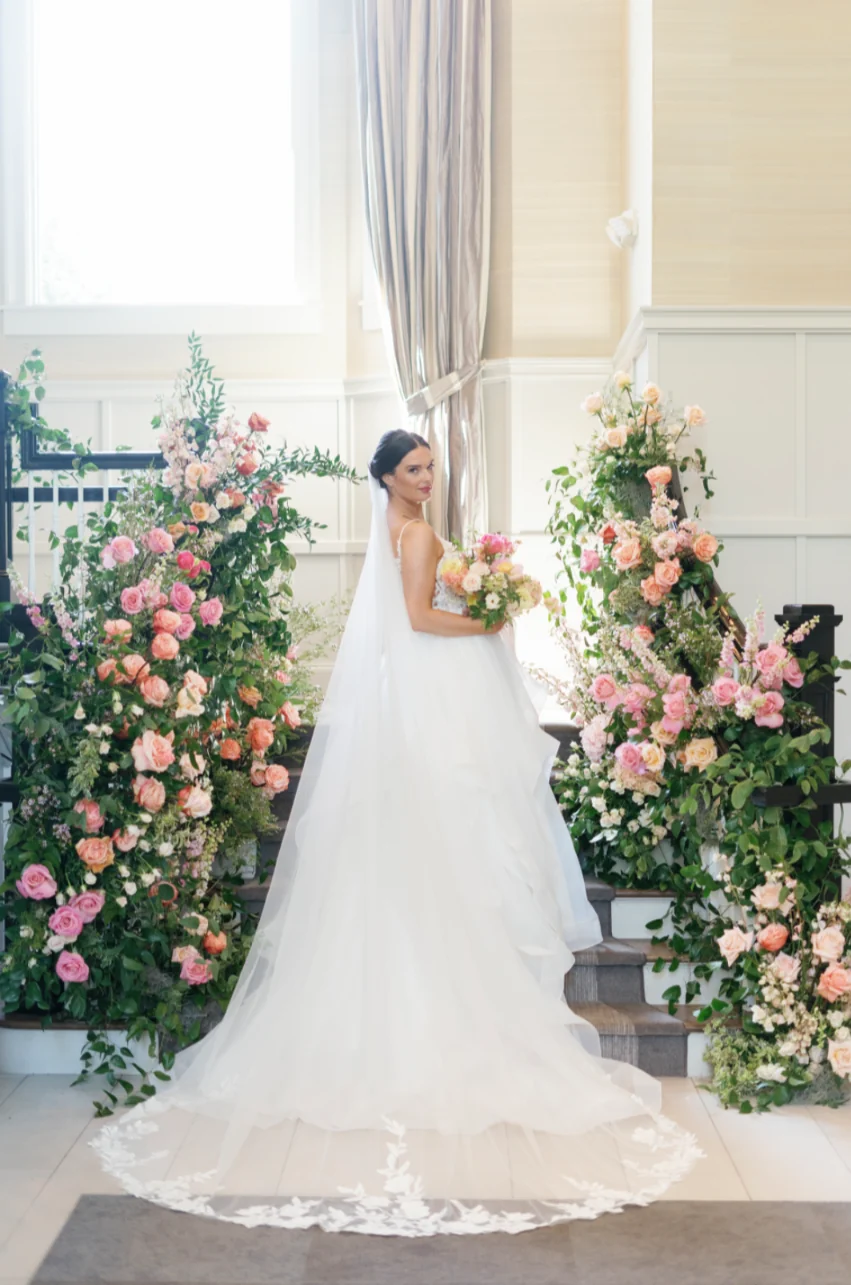 Bride on a staircase framed by two lush blush and coral floral installations