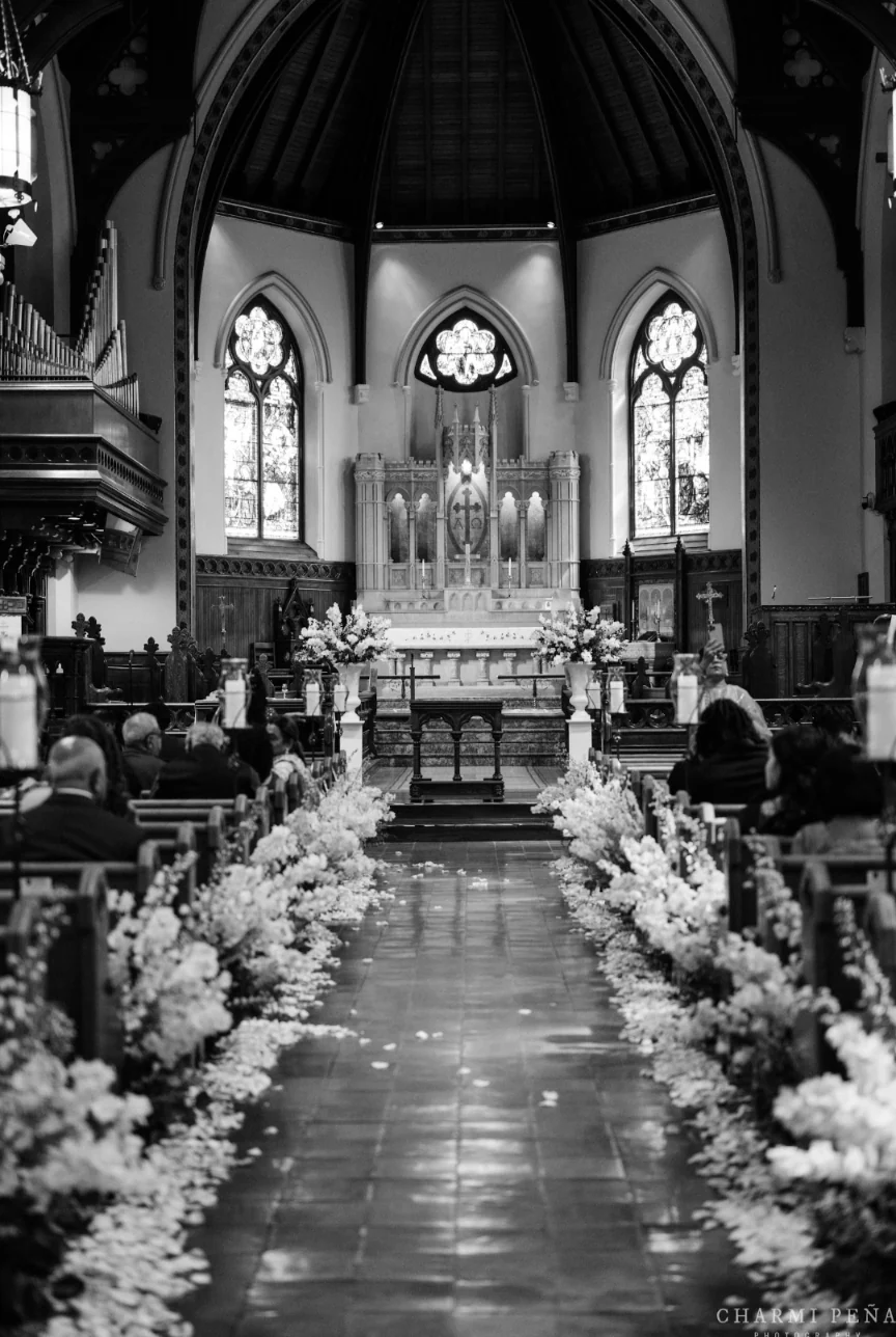 Church aisle lined with white floral arrangements and scattered petals, in black and white