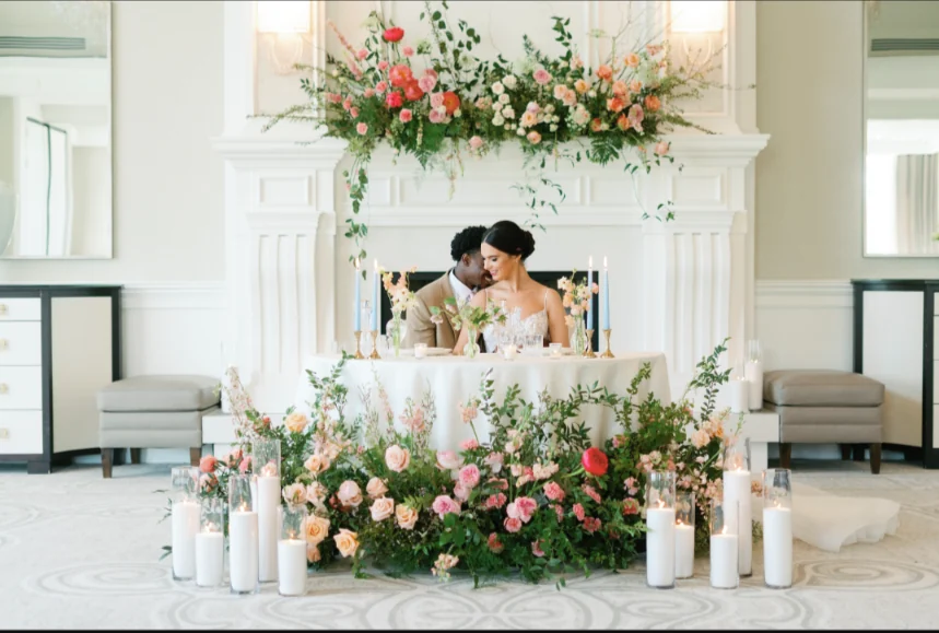 Sweetheart table with a low garden of peach, coral and blush roses and pillar candles, beneath a mantel arrangement